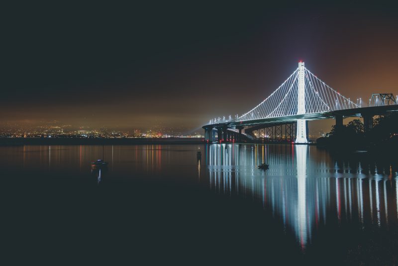 A suspension bridge lit at night, reflected in calm water.