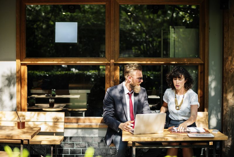 Two people working together at a laptop in a sunlit café.